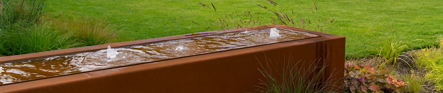 Een langwerpige, roestkleurige cortenstaal watertafel met meerdere kleine fonteintjes staat in een groene tuin met een strak gazon.