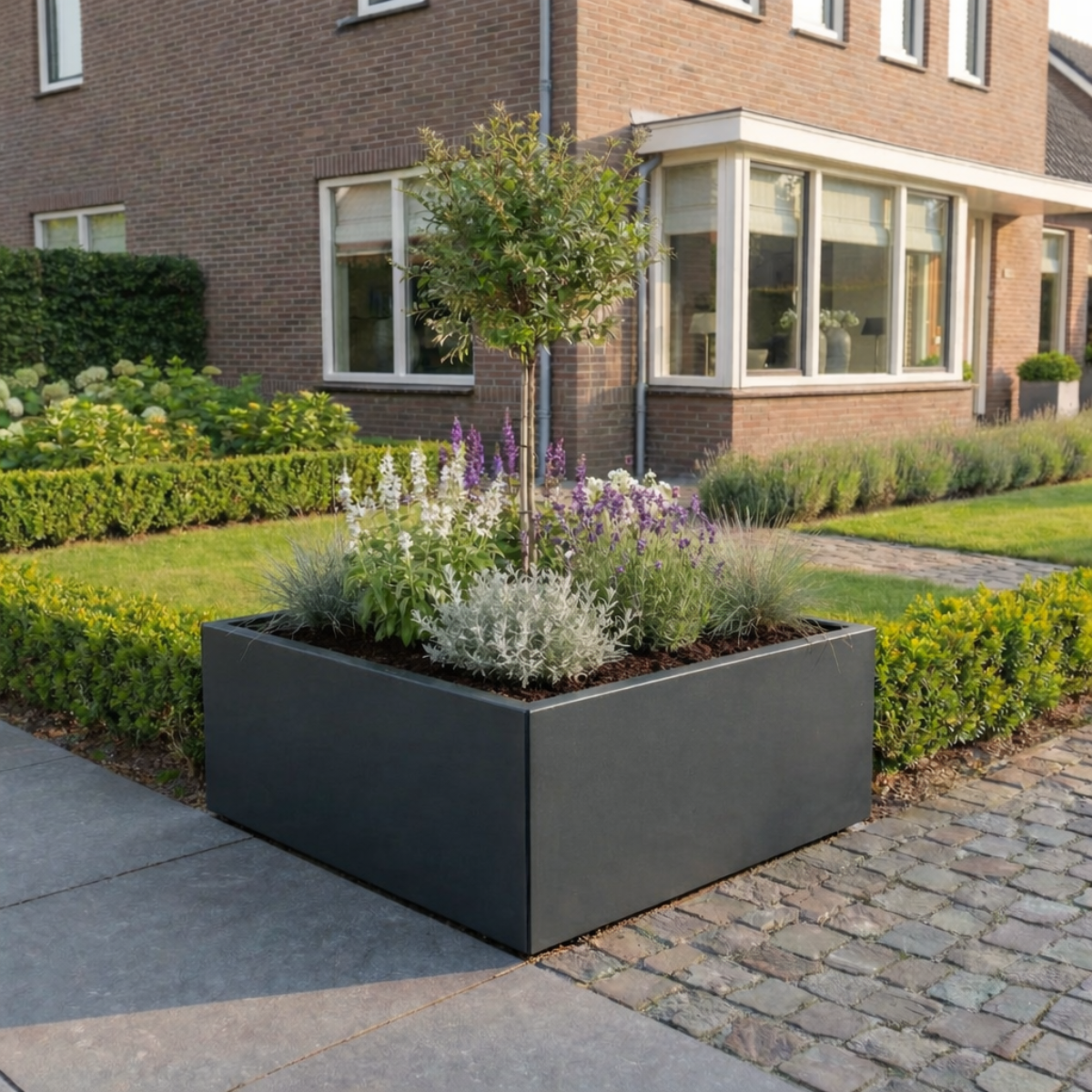 Vierkante, antracietgrijze plantenbak gevuld met een boompje op stam, witte en paarse bloemen in een moderne voortuin bij een bakstenen huis.