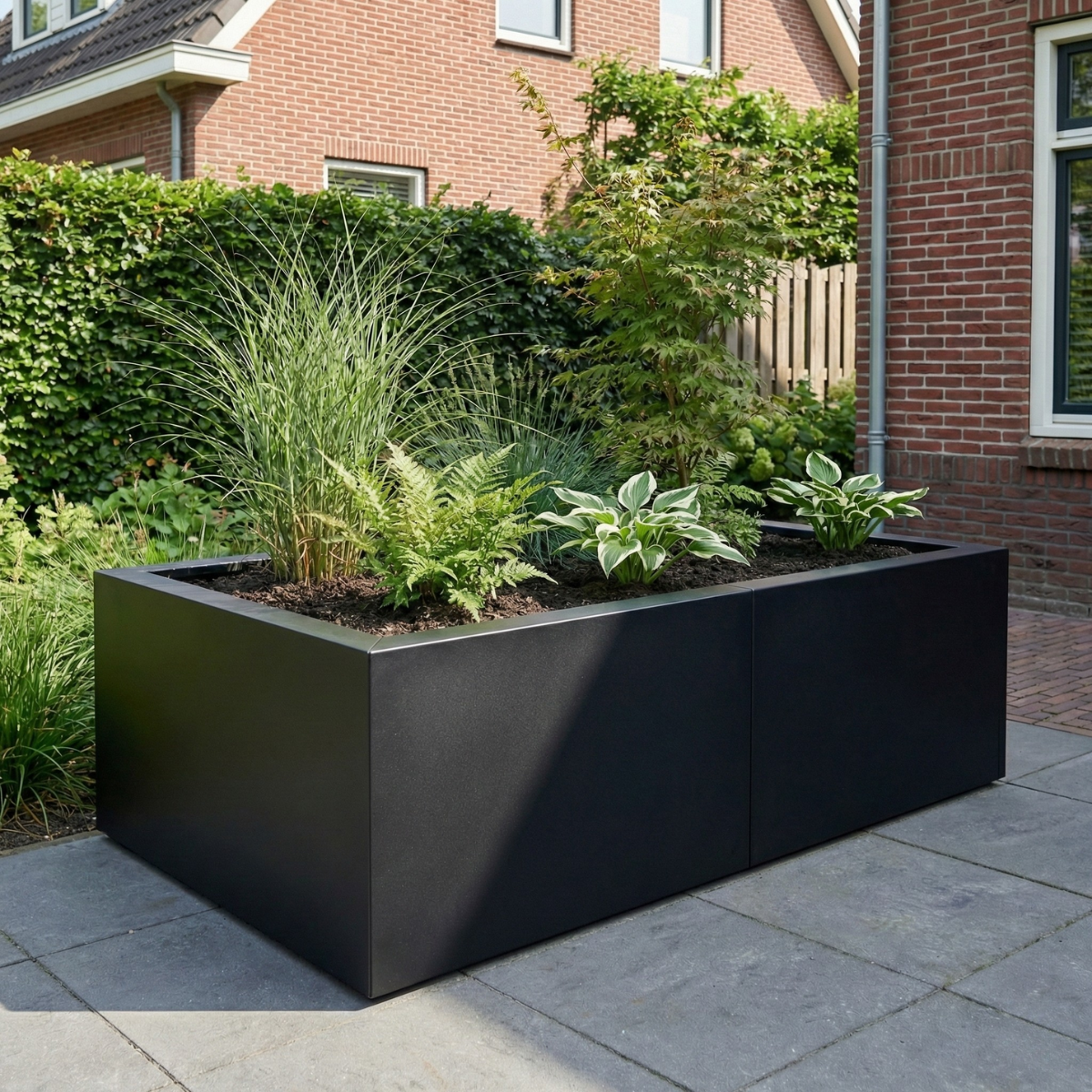Zwarte plantenbak gevuld met siergrassen en varens op een betegeld terras in een tuin.