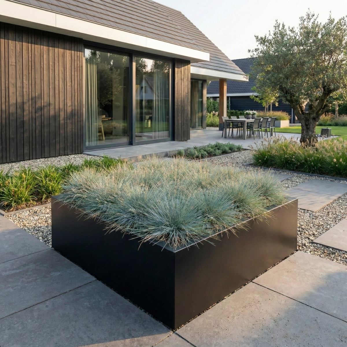 Grote zwarte plantenbak gevuld met blauwgrijs siergras in een moderne tuin met grind en grote tegels.