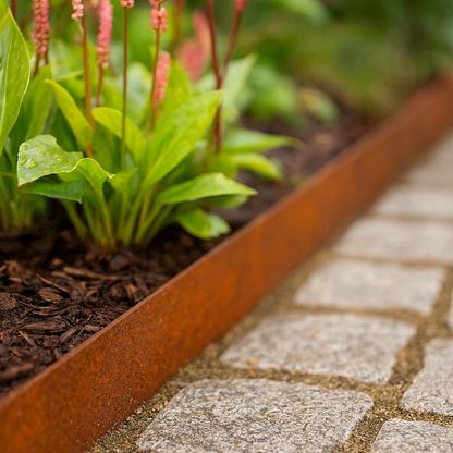Cortenstalen plantenbak gevuld met groene planten en roze bloemaren in een tuin met kasseien.