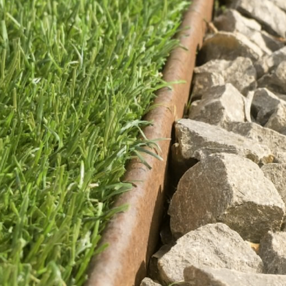Close-up van een cortenstalen plantenbak in een tuin, die een duidelijke scheiding vormt tussen een groen gazon en grote grijze stenen.