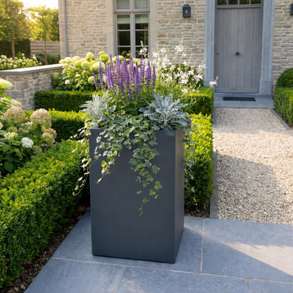 Hoge antracietgrijze plantenbak gevuld met paarse salvia en hangende klimop op een stenen terras in een weelderige voortuin met grind.