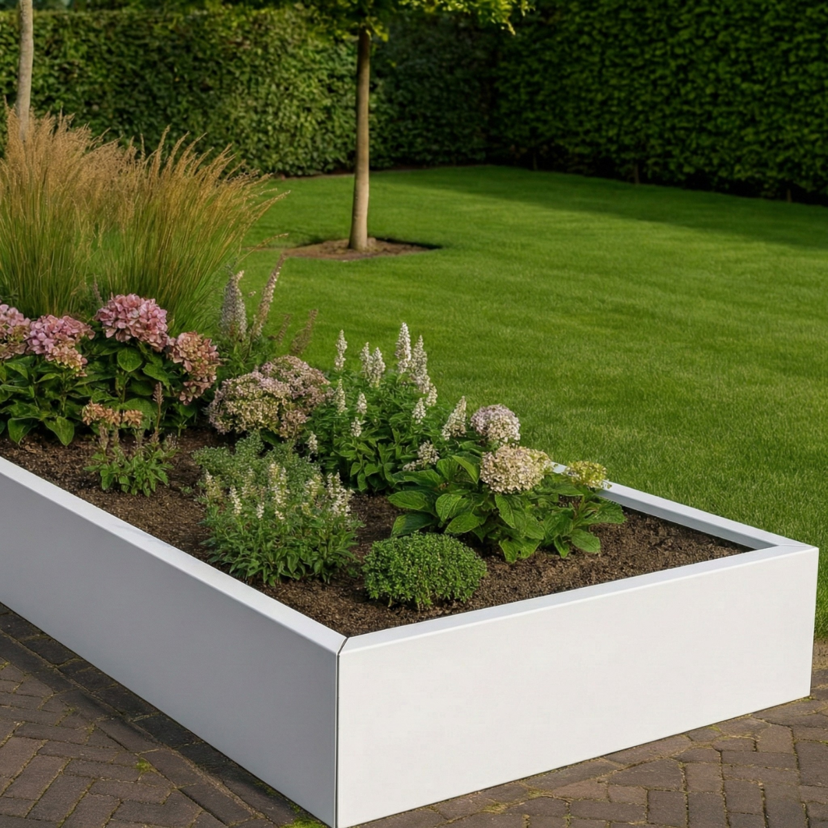 Grote witte plantenbak gevuld met roze hortensia's en siergrassen in een tuin met een gazon en klinkers.