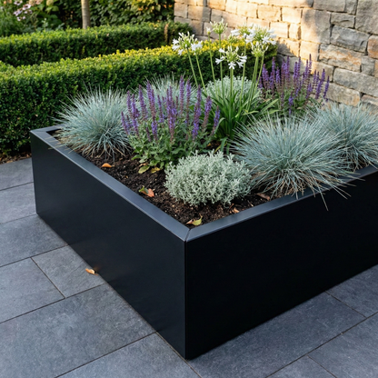 Grote zwarte rechthoekige plantenbak gevuld met siergrassen, paarse en witte bloemen op een terras met grijze tegels.