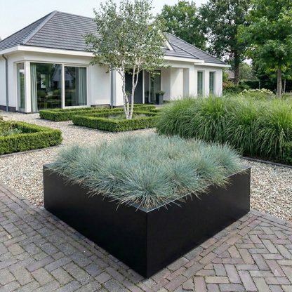 Grote zwarte plantenbak gevuld met siergrassen in een sfeervolle voortuin met grind bij een woning.