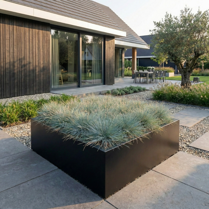 Grote zwarte plantenbak gevuld met blauwgrijs siergras in een moderne tuin met grind en grote tegels.