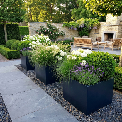 Drie donkergrijze vierkante plantenbakken ritmisch geplaatst langs een tuinpad, sfeervol gevuld met siergrassen, witte hortensia's, buxusbollen en paarse lavendel in een luxe tuin met buitenhaard.