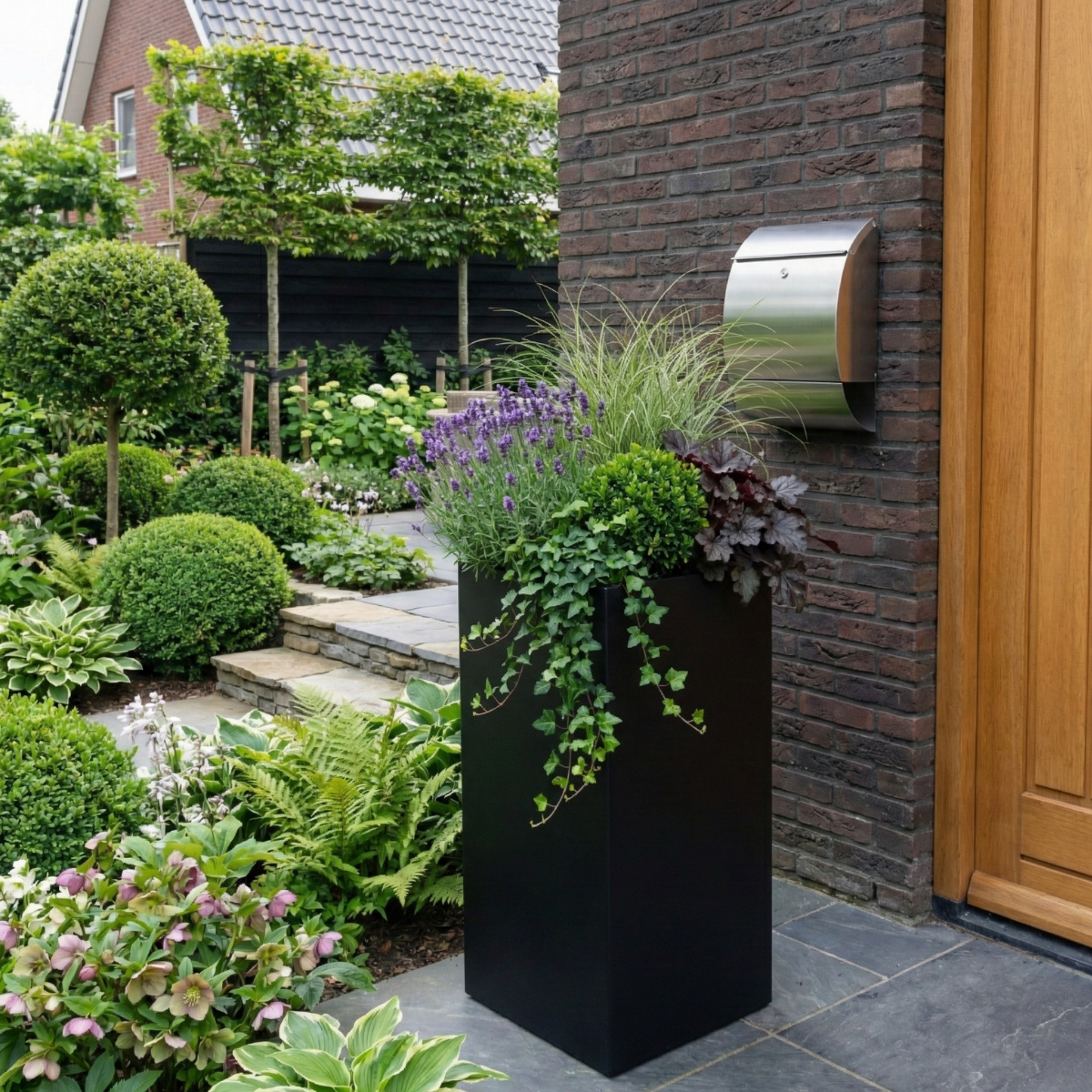 Hoge zwarte rechthoekige plantenbak gevuld met een mix van lavendel, siergrassen en hangende klimop, stijlvol geplaatst naast de voordeur onder een RVS brievenbus.