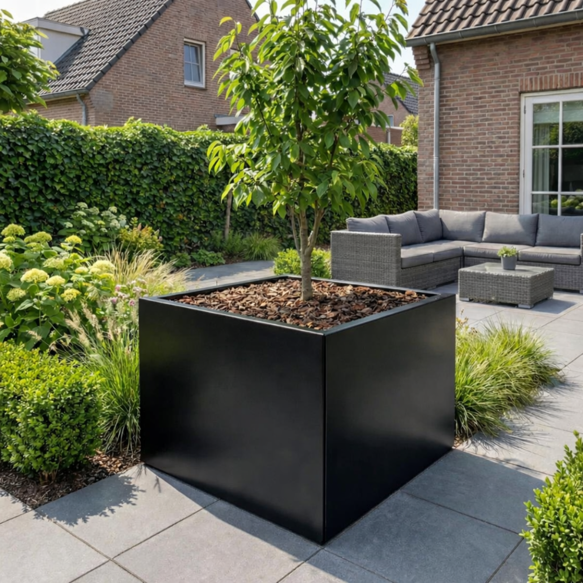 Vierkante zwarte plantenbak gevuld met een boompje en houtsnippers op een modern betegeld terras met een grijze loungeset op de achtergrond.